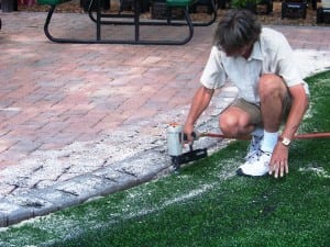 man stapling down artificial turf with sand infill near pavers