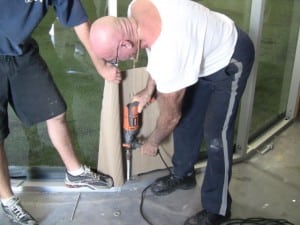 man drilling plexiglass wall into floor around indoor artificial turf field